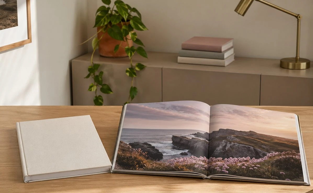 Aufgeschlagenes Fotobuch mit Küstenpanorama auf einem Holzschreibtisch, daneben ein geschlossenes Fotobuch im Leineneinband